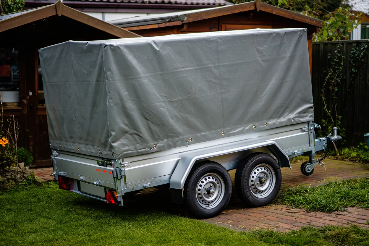 Remorque double essieux gris avec bâche PVC gris, avec roue de secours et timon, stationnée sur pavé devant jardin.