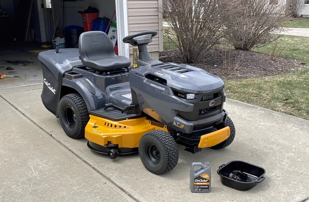 Tracteur tondeuse Cub Cadet garé devant un garage au printemps, avec un bac de vidange et une bouteille d'huile posés au sol pour l'entretien saisonnier.